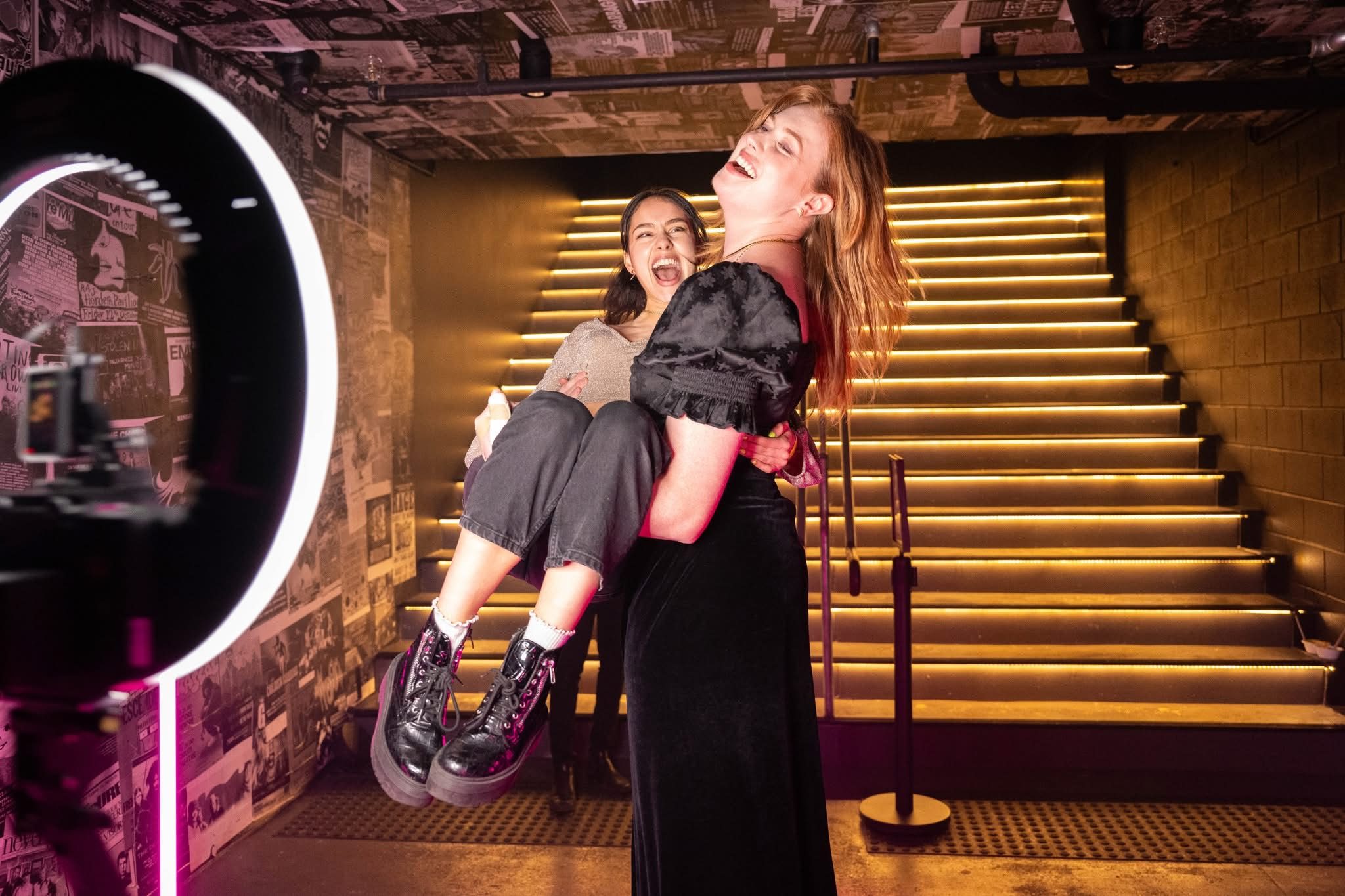A woman laughs as she carries her friend in front of glowing stairs